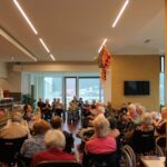 Več oseb sedi v skupnem prostoru, razporejene so v polkrogu in spremljajo dogajanje v sredini prostora.