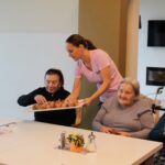 Oseba stoji ob mizi in ponuja pladenj z ocvrtimi kosi testa sedečim osebam.