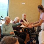 Oseba stoji ob skupini sedečih oseb in jim deli ocvrte kose testa s pladnja, več oseb jih že drži v rokah.