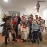 Skupina oseb v tradicionalnih kosmatih oblačilih z rogovi stoji v hodniku ob starejši osebi z hojco.