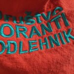 Bližnji posnetek rdečega naglavnega traku z izvezenim napisom »Društvo koranti Podlehnik«.