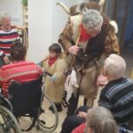 Oseba v kosmatem kostumu z rogovi stoji med sedečimi osebami v prostoru, ob njej je tudi otrok v podobnem kostumu.