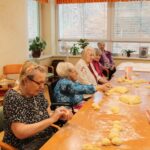 Več starejših oseb sedi za mizo in oblikuje kose testa.