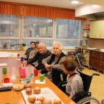 Skupina starejših oseb sedi za mizo s krofi in pijačo v prostoru s kuhinjskimi elementi.