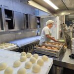 Oseba v kuhinji pripravlja krofe; na delovni površini so razporejeni kosi testa, poleg so pladnji z ocvrtimi krofi.