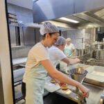 Oseba v kuhinji polni krof z nadevom s pomočjo ročnega polnilnika, v ozadju so drugi kuhinjski pripomočki.