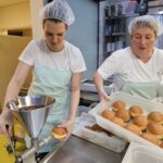 Dve osebi v kuhinji polnita in prestavljata krofe v plastične posode na delovni mizi.