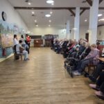 Skupina oseb stoji pred sedečim občinstvom, ena oseba igra kitaro, druge berejo besedilo.