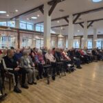 Več oseb sedi na stolih v dvorani in spremlja dogajanje pred seboj.