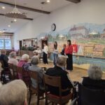 Skupina oseb stoji ob steni in nastopa pred sedečim občinstvom v notranjem prostoru.