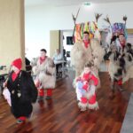 Več oseb v kosmatih pustnih kostumih in otroci v podobnih oblačilih hodijo po notranjem prostoru mimo sedečih oseb.