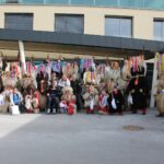 Skupina oseb v kosmatih pustnih kostumih in nekaj drugih oseb pozira pred vhodom v stavbo na prostem.