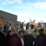 Osebe stojijo na prostem pred stavbo in opazujejo skupino oseb v kosmatih pustnih kostumih z barvnimi trakovi in perjem.