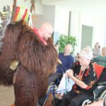 Oseba v kosmatem pustnem kostumu se sklanja k sedeči osebi v invalidskem vozičku, medtem ko druge osebe sedijo in opazujejo.