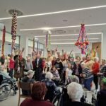 Skupina oseb stoji in sedi v notranjem prostoru, okrašenem z visečimi barvnimi trakovi, medtem ko jih druge osebe opazujejo z invalidskih vozičkov in stolov.