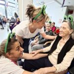 Več oseb z zelenimi naglavnimi dodatki sedi v prostoru, ena oseba pa se sklanja k sedeči osebi in jo drži za roko.