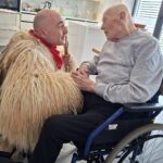 Oseba v invalidskem vozičku se pogovarja z drugo osebo v kosmatem pustnem kostumu, ki drži palico.