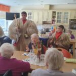 Kurenti v kožuhih in z zvonci stojijo ob mizi, za katero sedijo stanovalci v jedilnici.