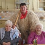 Kurent stoji za dvema nasmejanima stanovalkama, ki sedita za mizo v skupnem prostoru.