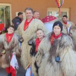 Mlajši in starejši kurenti pozirajo pred domom; v ozadju visi pisan pustni okras.