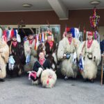 Skupina oseb v tradicionalnih kožuhastih pustnih kostumih stoji pred vhodom v stavbo.