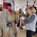 Oseba v kožuhastem pustnem kostumu stoji pred žensko, ki na palico privezuje trakove blaga.