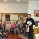 Oseba stoji pred sedečimi osebami v notranjem prostoru in v rokah drži barvit papirnat okras.