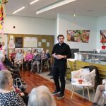 Oseba stoji v prostoru in govori zbranim sedečim osebam, medtem ko druga oseba sedi ob strani.