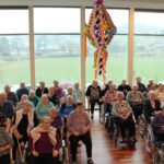 Skupina oseb sedi na stolih in v invalidskih vozičkih v svetlem prostoru z velikimi okni in visečim barvnim okrasom.