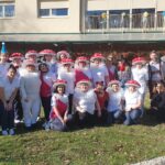 Skupina odraslih stoji na travi pred stavbo; večina je oblečena v bele kostume z rdečimi pikastimi klobuki v obliki mušnic.