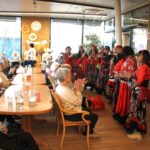 Skupina izvajalk v rdeče-črnih kostumih nastopa pred sedečimi stanovalci v skupnem prostoru.