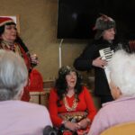 Izvajalka v rdečem kostumu stoji ob harmonikarju, medtem ko stanovalci sedijo pred njima.