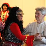 Izvajalka v rdeče-črnem kostumu pleše s stanovalko v svetlem puloverju.