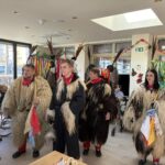 Skupina kurentov v tradicionalnih kožuhih in z okrašenimi pokrivali stoji v svetlem prostoru doma in obiskuje stanovalce.