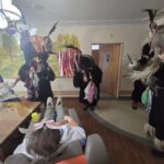 Kurenti v črnih kožuhih z zvonci nastopajo v sobi, medtem ko stanovalci sedijo za mizo in jih opazujejo.