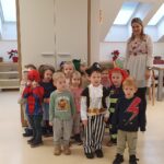 Skupina otrok v pustnih kostumih stoji v prostoru, za njimi stoji vzgojiteljica.