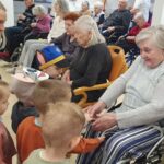 Otroci stojijo ob starejši ženski na vozičku in se z njo pogovarjajo ter držijo za roke.