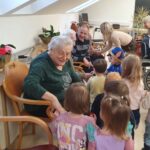 Starejša ženska sedi na stolu in se pogovarja z otroki, ki stojijo okoli nje.