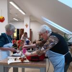 Dve starejši ženski za mizo delita pecivo, v ozadju so otroci in odrasli.