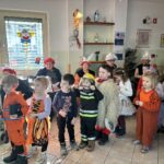 Skupina otrok v pustnih kostumih stoji v prostoru, medtem ko za njimi sedijo starejše osebe ob oknu.