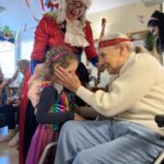 Starejša oseba sedi na stolu in se pogovarja z otrokom v pustnem kostumu, ob njiju stoji oseba v barvitem kostumu.