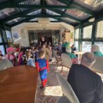 Otroci v pustnih kostumih stojijo in se pogovarjajo s sedečimi starejšimi osebami v svetlem notranjem prostoru.