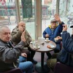 Štirje moški sedijo za mizo ob oknu, pijejo kavo in mahajo proti fotoaparatu.