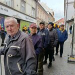 Skupina moških stoji ob ograji na mestni ulici in opazuje dogajanje.