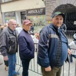 Trije moški stojijo ob kovinski ograji na ulici; v ozadju so trgovine in mimoidoči.