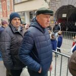 Moški v zimskih oblačilih stojijo ob ograji in gledajo proti ulici, kjer poteka dogajanje.