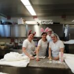 Štiri osebe stojijo za delovno mizo v kuhinji, nekatere z naglavnimi dodatki, na mizi pa je testo in kuhinjski pripomočki.