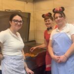 Tri osebe stojijo ob cvrtniku v kuhinji, ena oseba nosi pustni naglavni dodatek, v napravi pa so krofi.