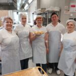 Pet oseb v kuhinji stoji skupaj, ena oseba drži krožnik s krofi.