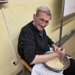 Moški sedi na stolu in z rokami meša testo v kovinski posodi.